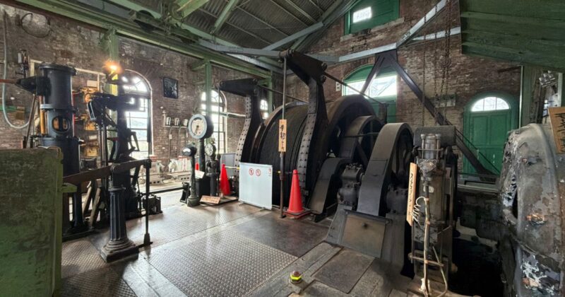 万田坑の巻上機と操作レバー。隣り合うアナログな計器と巨大な鉄製機械が並ぶ機械室の風景。