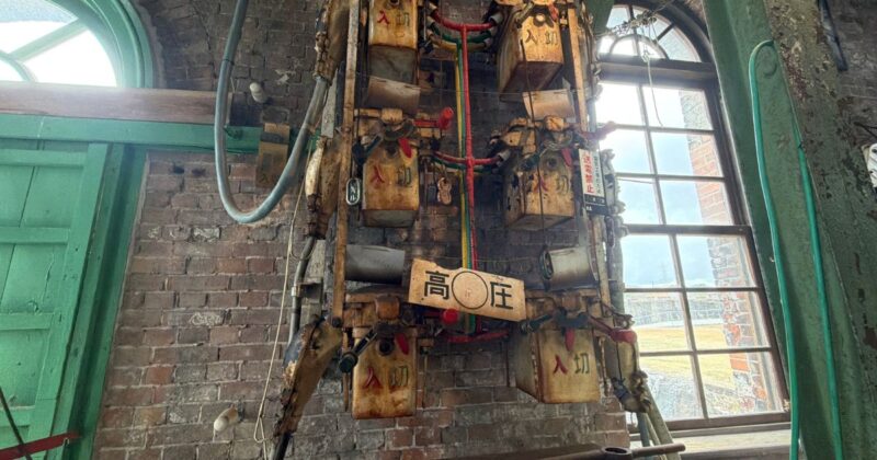万田坑機械室の壁に設置された古い高圧電気のスイッチ。錆びた質感と「入」「切」の文字、レンガ壁が織りなすレトロな風景。