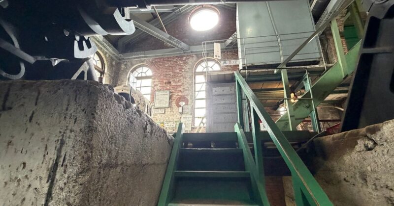 万田坑の機械室にある緑色の鉄製階段と赤いレンガ壁。天井の窓から光が差し込むレトロな産業遺産の風景。